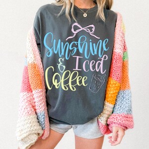 Op de afbeelding: Donkergrijze sweatshirt met de tekst "Sunshine & Iced Coffee" in blauw, roze en geel. Een kleurrijk patchwork-cardigan in roze, oranje, geel en blauw wordt over de sweatshirt gedragen. De sweatshirt heeft een strik-afbeelding.
