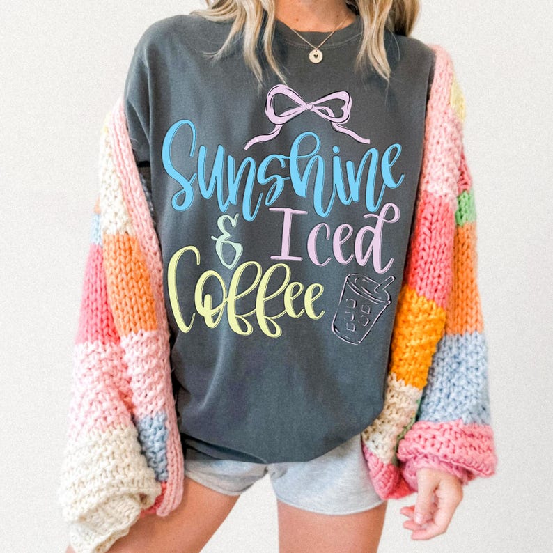 Op de afbeelding: Donkergrijze sweatshirt met de tekst "Sunshine & Iced Coffee" in blauw, roze en geel. Een kleurrijk patchwork-cardigan in roze, oranje, geel en blauw wordt over de sweatshirt gedragen. De sweatshirt heeft een strik-afbeelding.