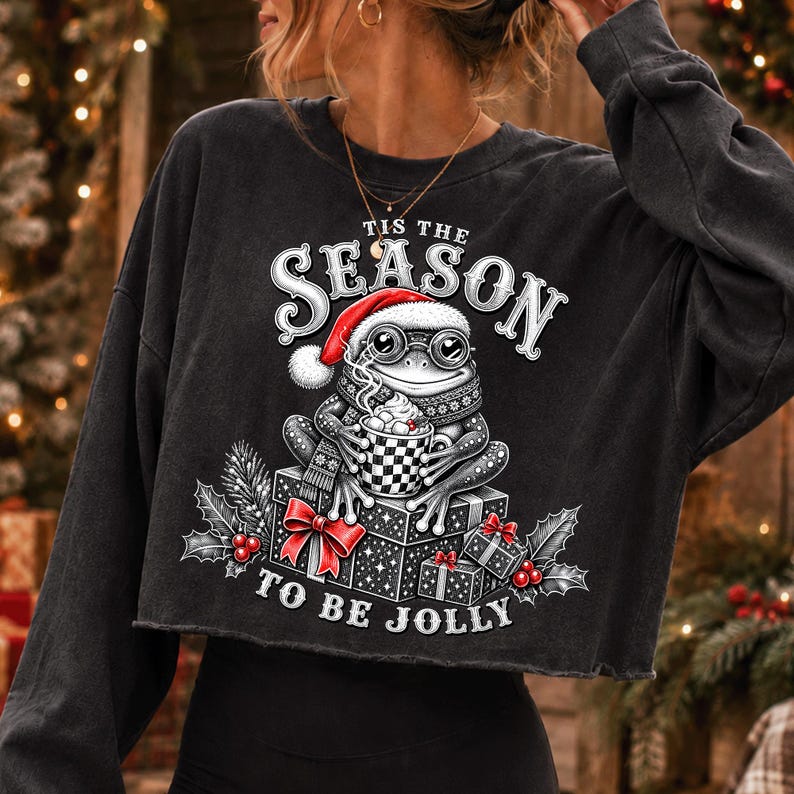 Puede incluir: Una sudadera negra de manga larga y corte corto con el gr&aacute;fico de una rana con gorro de Pap&aacute; Noel, bufanda y gafas, sosteniendo una taza. El texto Tis The Season To Be Jolly est&aacute; rodeado de cajas de regalo, hojas de acebo y bayas rojas.