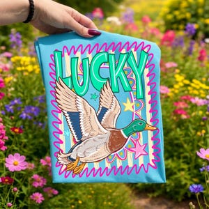 May include: A light blue t-shirt with a cartoon duck in flight and the word "LUCKY" in green bubble letters. The duck has brown, white, and blue wings, a green head, and an orange beak. Pink and yellow stars and wavy lines surround the duck.