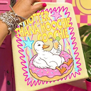 May include: A light yellow graphic tee featuring a cartoon duck relaxing on a pink donut float and holding a cocktail. The text "OTTER THANA HOOCHIE COOCHIE" is displayed in a colorful, wavy font. The design includes stars and a wavy border.