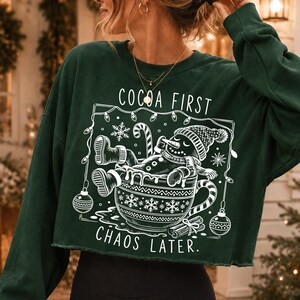 Op de afbeelding: Een donkergroene, langarmige cropped sweater met een witte afbeelding van een sneeuwpop die ontspant in een grote mok. De tekst luidt COCOA FIRST CHAOS LATER. Het ontwerp bevat sneeuwvlokken, een zuurstok en kerstballen in een vierkant kader.
