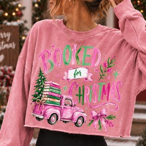 May include: A dusty rose long-sleeve sweatshirt featuring a graphic print with the text "BOOKED for CHRISTMAS". The design includes a pink pickup truck carrying a stack of books and a Christmas tree, accented by a pink bow and green foliage.