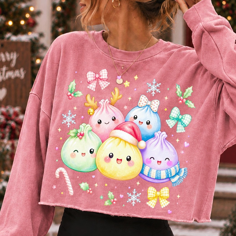 May include: A dusty rose long-sleeve cropped sweatshirt featuring a festive graphic of five colourful, smiling dumpling characters. The dumplings are decorated with holiday elements including a Santa hat, a scarf, reindeer antlers, and bows.
