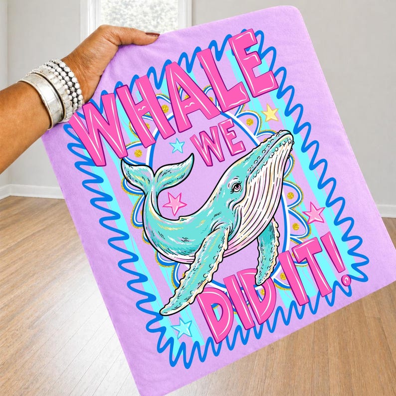 K&ouml;nnte beinhalten: Eine hellviolette Decke mit einer Cartoon-Wal-Illustration. Der Wal ist hellblau mit wei&szlig;en Details. Der Text "WHALE WE DID IT!" ist rosa, wei&szlig; umrandet. Die Decke wird von einer Person mit silbernen Armb&auml;ndern gehalten.