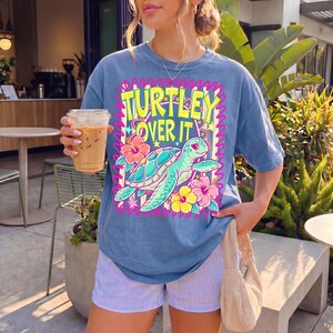 Puede incluir: Camiseta azul con el texto "TURTLEY OVER IT" en amarillo y rosa. La camiseta presenta una tortuga de dibujos animados con flores de hibisco. La persona lleva pantalones cortos a rayas azules y blancas y sostiene una bebida.