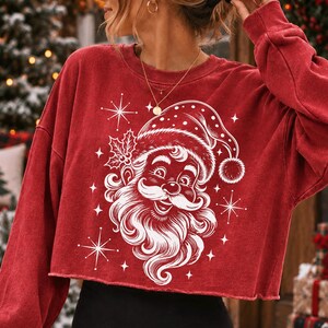 May include: A red long-sleeve cropped sweatshirt featuring a white graphic print of Santa Claus wearing a hat with holly leaves. The design includes several four-pointed starbursts surrounding the central portrait of Santa with a curly beard.