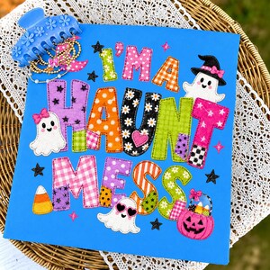 May include: A bright blue square sign featuring the colorful appliqued text I'M A HAUNT MESS with various patterns like polka dots, gingham, and daisies. The design includes three white ghosts, a pink jack-o'-lantern bucket, and black stars.