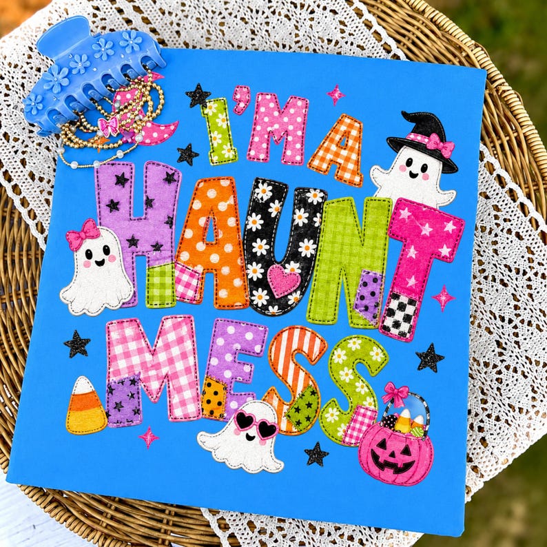 May include: A bright blue square sign featuring the colorful appliqued text I'M A HAUNT MESS with various patterns like polka dots, gingham, and daisies. The design includes three white ghosts, a pink jack-o'-lantern bucket, and black stars.