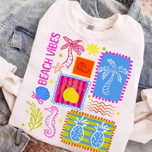 May include: White sweatshirt with a colourful "Beach Vibes" graphic. The design features stamps with palm trees, a starfish, a seashell, a seahorse, and pineapples. The text "Beach Vibes" is in pink. The sweatshirt is displayed on a denim jacket.