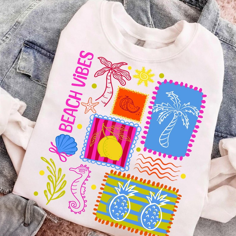 May include: White sweatshirt with a colourful "Beach Vibes" graphic. The design features stamps with palm trees, a starfish, a seashell, a seahorse, and pineapples. The text "Beach Vibes" is in pink. The sweatshirt is displayed on a denim jacket.