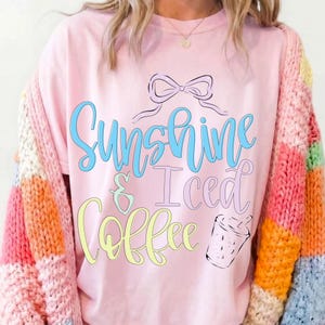 Op de afbeelding: Roze T-shirt met de tekst "Sunshine & Iced Coffee" in blauwe, gele en roze letters. Een zwarte strik en een tekening van een koffiekopje staan boven en rechts van de tekst. De persoon draagt een kleurrijke trui.