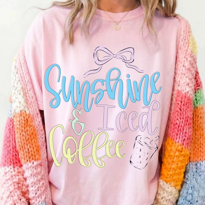 Op de afbeelding: Roze T-shirt met de tekst "Sunshine & Iced Coffee" in blauwe, gele en roze letters. Een zwarte strik en een tekening van een koffiekopje staan boven en rechts van de tekst. De persoon draagt een kleurrijke trui.
