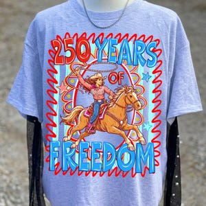 May include: A light blue t-shirt featuring a graphic of a cowgirl riding a horse, roping a lasso. The text reads "250 Years of Freedom" in red and blue. The design is framed by a red zigzag border.