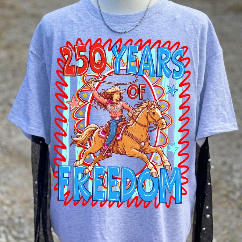May include: A light blue t-shirt featuring a graphic of a cowgirl riding a horse, roping a lasso. The text reads "250 Years of Freedom" in red and blue. The design is framed by a red zigzag border.