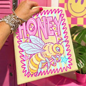 Pode incluir: Uma tela amarela com a palavra "HONEY" em letras rosa e uma ilustração de abelha de desenho animado. A abelha é amarela e laranja com asas e detalhes azuis. O design é emoldurado por uma borda ondulada rosa e estrelas.