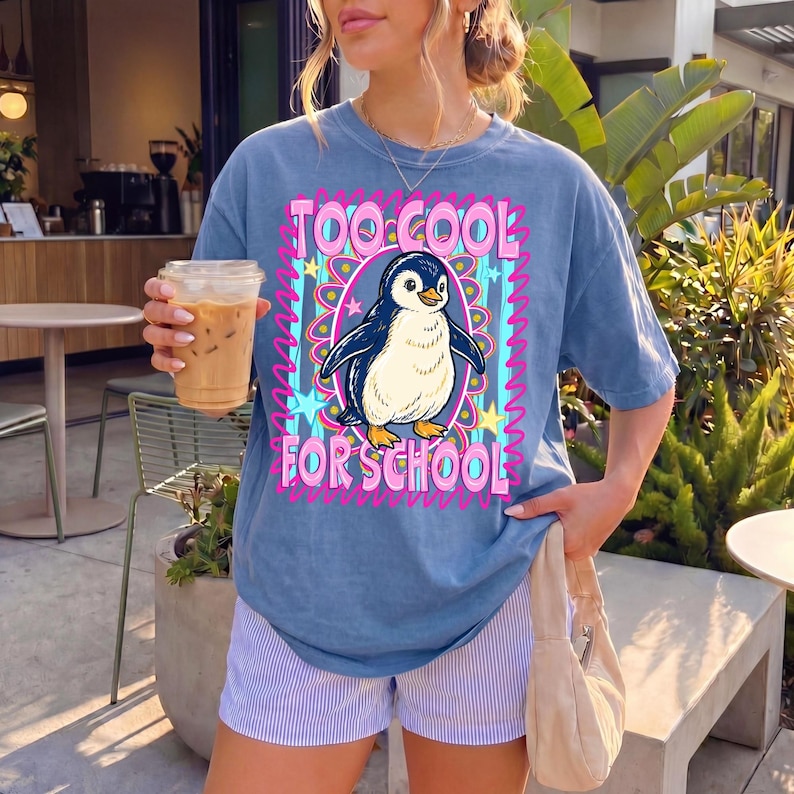 K&ouml;nnte beinhalten: Blaues T-Shirt mit einem Pinguin-Motiv in einem rosa und blauen Rahmen. Der Text "TOO COOL FOR SCHOOL" ist zu sehen. Das Shirt wird mit gestreiften Shorts getragen.