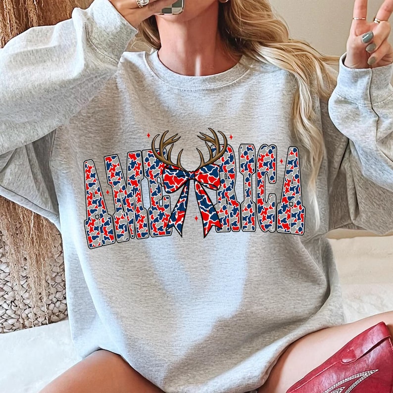 Pu&ograve; includere: Una felpa grigio chiaro con la scritta "AMERICA" in un design patriottico. Le lettere sono riempite con un motivo mimetico rosso, bianco e blu. Un fiocco con corna &egrave; centrato sopra la parola, aggiungendo un tocco unico.