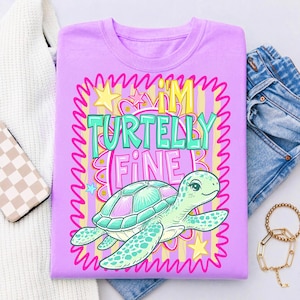 May include: A lilac t-shirt with a cartoon turtle graphic. The turtle is teal and pink, with the words "I'M TURTELLY FINE" in teal and pink. The design is framed with a pink, yellow, and teal border. The shirt is folded with a pair of jeans and a sweater.