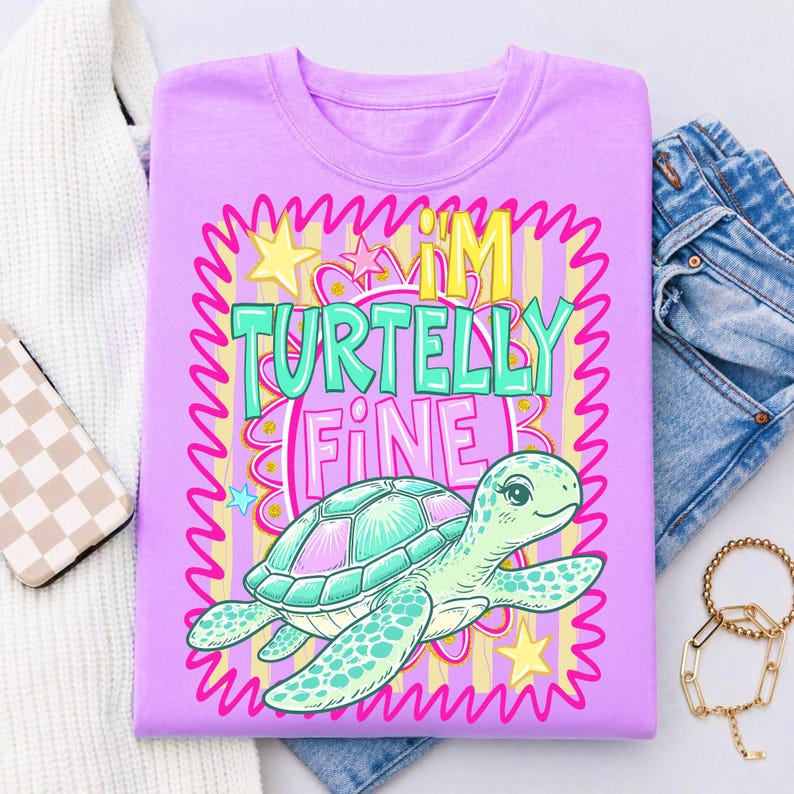 May include: A lilac t-shirt with a cartoon turtle graphic. The turtle is teal and pink, with the words "I'M TURTELLY FINE" in teal and pink. The design is framed with a pink, yellow, and teal border. The shirt is folded with a pair of jeans and a sweater.
