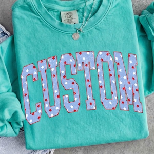 Op de afbeelding: Teal Comfort Colors sweatshirt met het woord "CUSTOM" in grote, versleten letters. De letters zijn lichtblauw met witte sterren en rode contouren. Een zilveren ketting met een hanger is zichtbaar.