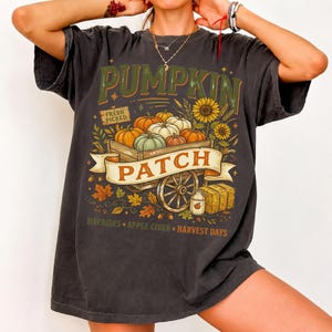 Puede incluir: Una camiseta gris carb&oacute;n de manga corta con un gr&aacute;fico de un carro de madera lleno de calabazas. El texto dice PUMPKIN PATCH, FRESH PICKED, HAYRIDES, APPLE CIDER y HARVEST DAYS. El dise&ntilde;o incluye girasoles, hojas de oto&ntilde;o y balas de heno.
