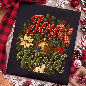 Może przedstawiać: Czarna koszulka z kolorową grafiką i napisem Joy to the World. Wz&oacute;r przedstawia scenę narodzin, białą gwiazdę betlejemską, czerwone jagody, zielone gałązki sosny, kokardę w kratę oraz małe jagnię odpoczywające u dołu nadruku.