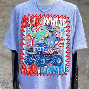 May include: Light blue t-shirt featuring a graphic of a green dinosaur wearing sunglasses, atop a monster truck with an American flag. The text reads "RED WHITE AND RAWR!" in red and white.
