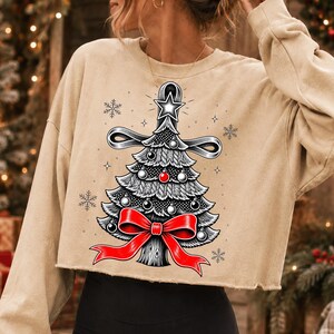 K&ouml;nnte beinhalten: Ein beigefarbenes, lang&auml;rmeliges bauchfreies Sweatshirt mit einer grafischen Illustration eines stilisierten Weihnachtsbaums. Der Baum ist mit einer gro&szlig;en roten Schleife an der Basis, einem Stern an der Spitze und kleinen Kugeln verziert.