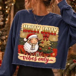 Może przedstawiać: Granatowa bluza z długim rękawem z nadrukiem przedstawiającym owcę w czapce Mikołaja i szaliku obok udekorowanej choinki. Napis HOLLY JOLLY znajduje się u g&oacute;ry, a napis vibes only czerwoną czcionką znajduje się na dole grafiki.