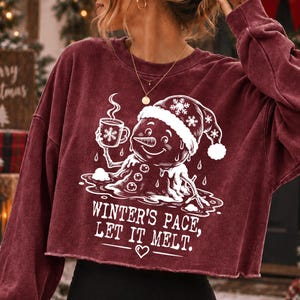 May include: A maroon long-sleeve cropped sweatshirt featuring a white graphic of a melting snowman wearing a Santa hat and holding a mug. The text reads "WINTER'S PACE, LET IT MELT." with a small heart icon below the lettering on the front.