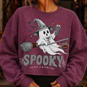 May include: A dark maroon long-sleeve sweatshirt featuring a graphic of a ghost wearing a witch hat, riding a broomstick, and holding a drink. The ghost is accompanied by bats, a pumpkin, and the text SPOOKY HAPPY HAUNTING in a checkered pattern.