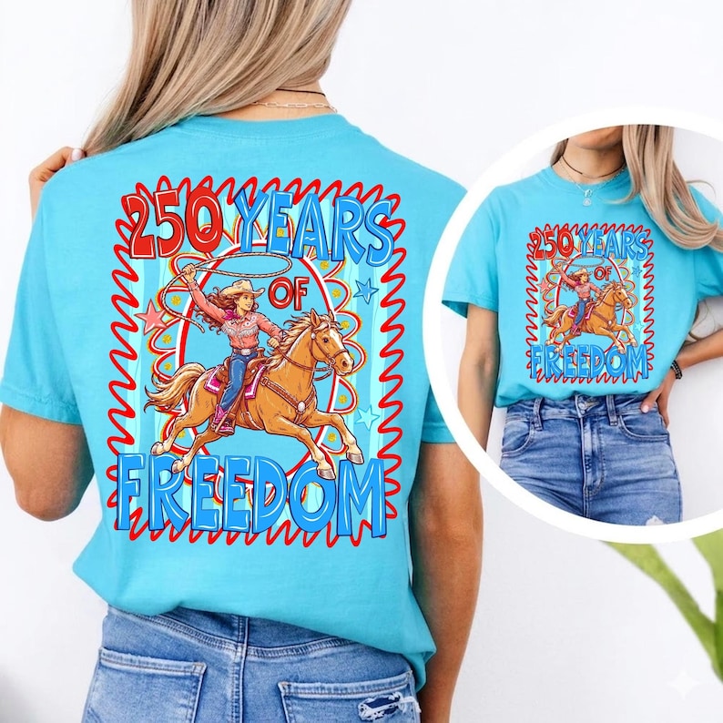May include: A light blue t-shirt featuring a graphic of a woman riding a horse, roping a steer, with the text "250 Years of Freedom". The design is framed with a red and blue wavy border.