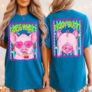 Op de afbeelding: Teal t-shirt met een cartoon varken met een hartvormige zonnebril en kersen. De voorkant zegt "GUESS WHAT?" en de achterkant zegt "PIGGY BUTT!" in een neon-stijl lettertype. De varkensafbeelding is omlijnd in roze.