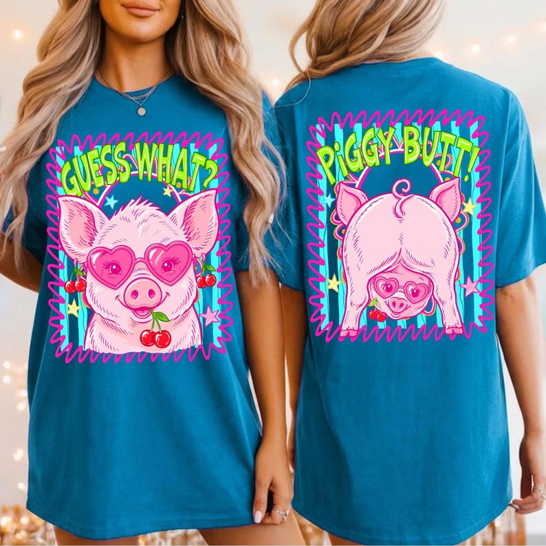 Op de afbeelding: Teal t-shirt met een cartoon varken met een hartvormige zonnebril en kersen. De voorkant zegt "GUESS WHAT?" en de achterkant zegt "PIGGY BUTT!" in een neon-stijl lettertype. De varkensafbeelding is omlijnd in roze.