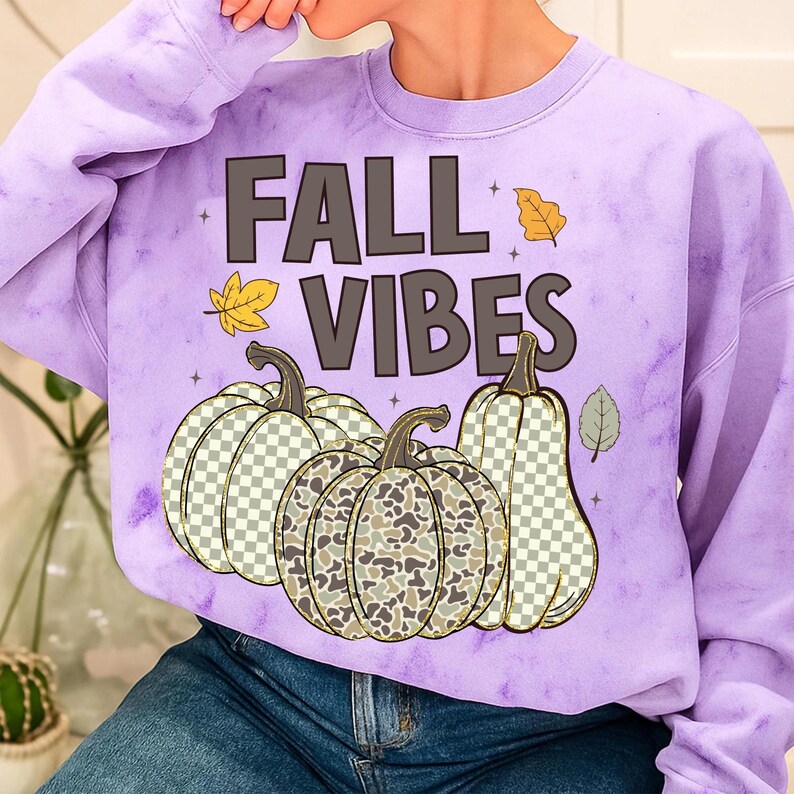 Puede incluir: Una sudadera tie-dye de color morado claro con el texto FALL VIBES en letras de color marr&oacute;n oscuro. El dise&ntilde;o incluye tres calabazas con patrones variados, incluyendo un estampado de cuadros y uno de camuflaje, con hojas amarillas y verdes.