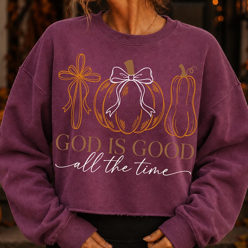 Puede incluir: Una sudadera de manga larga de color morado oscuro con un gr&aacute;fico de una cruz, una calabaza con un lazo blanco y una calabaza alargada. El texto "GOD IS GOOD all the time" est&aacute; impreso en letras doradas y blancas en el centro.