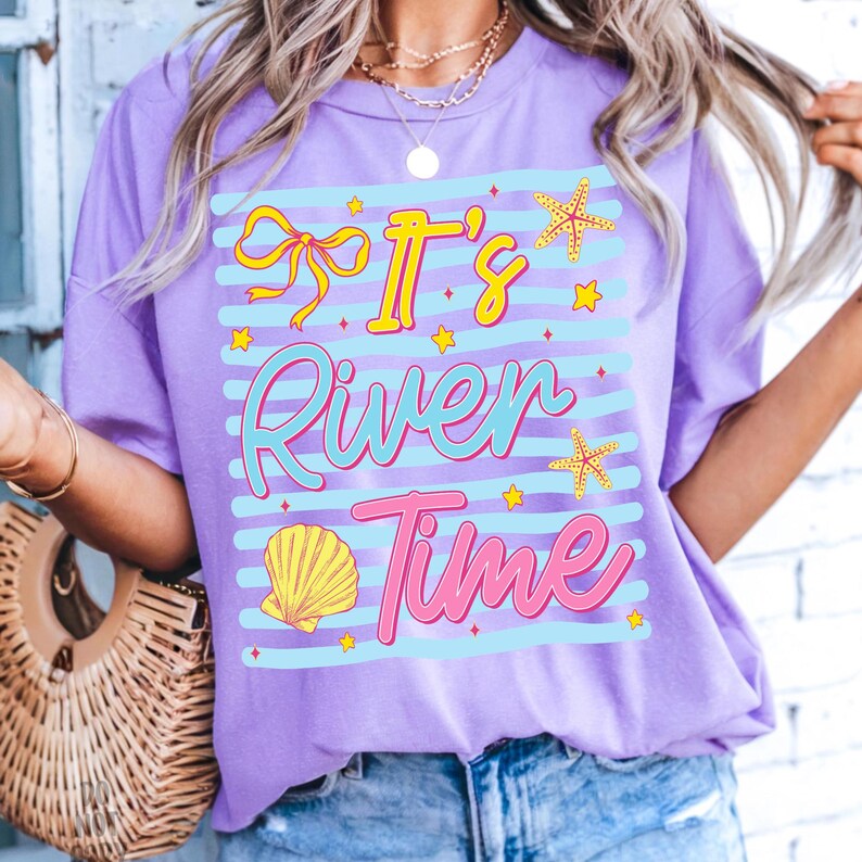 May include: A lilac-colored t-shirt with the text "It's River Time" in a playful, colorful design. The design includes a bow, starfish, a seashell, and stars. The shirt has a relaxed fit and short sleeves.
