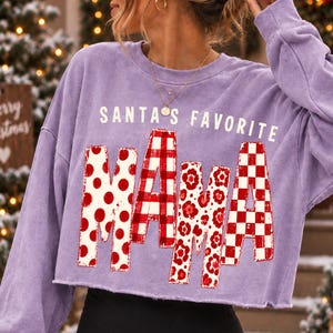 K&ouml;nnte beinhalten: Ein hellviolettes, lang&auml;rmeliges Cropped-Sweatshirt mit dem wei&szlig;en Schriftzug SANTA'S FAVORITE &uuml;ber dem Wort MAMA in gro&szlig;en Blockbuchstaben. Jeder Buchstabe von MAMA zeigt ein anderes rot-wei&szlig;es Muster, darunter Punkte, Karos und Blumen.