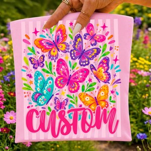 May include: A light pink fabric pouch featuring a vertical striped pattern and a colourful graphic print of various butterflies in shades of orange, purple, pink, and teal, with the word CUSTOM written in large, bold, bright pink script at the bottom.