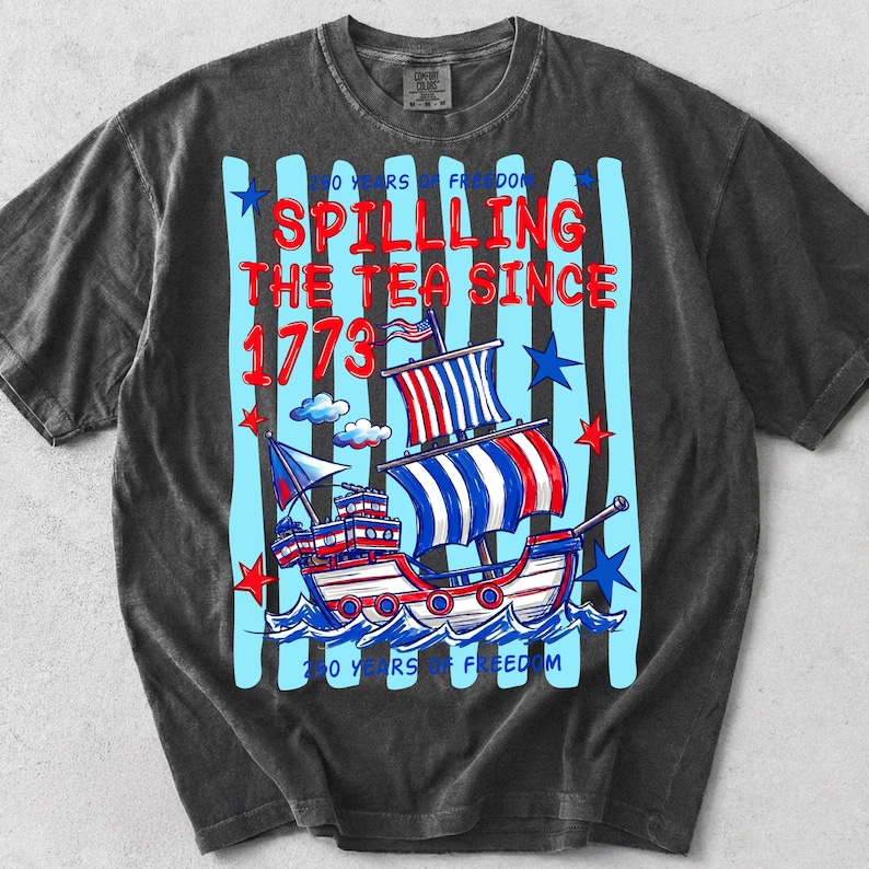 Pu&ograve; includere: T-shirt grigio scuro con un design grafico raffigurante una nave rossa, bianca e blu che naviga sulle onde. Il testo "Spilling the Tea Since 1773" &egrave; in rosso, con strisce verticali blu e stelle. La maglietta ha anche il testo "250 Years of Freedom".