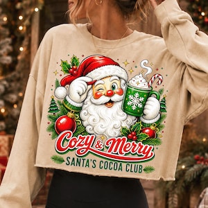 K&ouml;nnte beinhalten: Ein beiges Langarm-Sweatshirt mit einer Grafik eines zwinkernden Weihnachtsmanns, der eine gr&uuml;ne Tasse mit Schneeflockenmuster und einer Zuckerstange h&auml;lt. Der Text lautet Cozy & Merry SANTA'S COCOA CLUB, umgeben von roten Kugeln und Tannenzweigen.