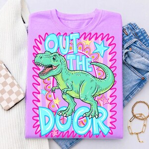 May include: A light purple t-shirt featuring a cartoon Tyrannosaurus Rex in shades of green. The text "OUT THE DOOR" is displayed in a bubble font. The design includes pink and blue accents, stars, and wavy lines.