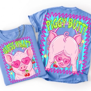 Op de afbeelding: Twee lichtblauwe t-shirts met een cartoon varkensontwerp. E&eacute;n shirt heeft een varken met een hartvormige zonnebril en de tekst "GUESS WHAT?". Het andere shirt toont de achterkant van het varken met de tekst "PIGGY BUTT!".