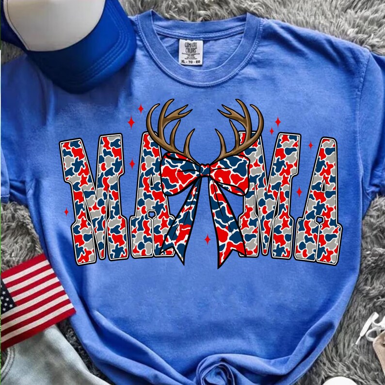 Pu&ograve; includere: Una maglietta blu con la scritta "MAMA" in un motivo mimetico rosso, bianco e blu. Un fiocco con corna di cervo &egrave; centrato sopra la parola. Accenti di stelle rosse sono posizionati attorno al testo. Un berretto da baseball blu e una bandiera americana sono sullo sfondo.