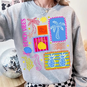 May include: A grey sweatshirt with the words "Beach Vibes" in pink. The sweatshirt features colourful graphic stamps with palm trees, a lemon, a seahorse, and pineapples. The design includes a seashell, starfish, and sun.