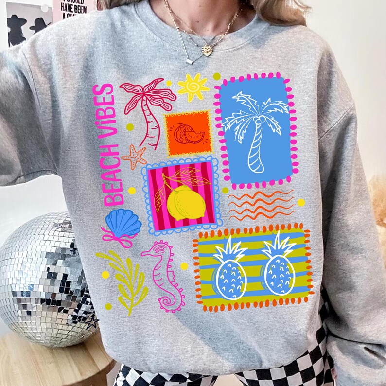 May include: A grey sweatshirt with the words "Beach Vibes" in pink. The sweatshirt features colourful graphic stamps with palm trees, a lemon, a seahorse, and pineapples. The design includes a seashell, starfish, and sun.