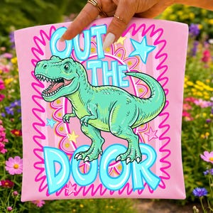 May include: Pink t-shirt with a turquoise Tyrannosaurus Rex illustration and the text "OUT THE DOOR" in turquoise. The design includes stars and a wavy border.