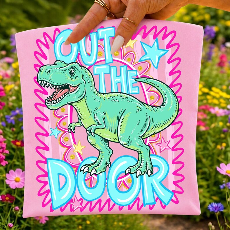May include: Pink t-shirt with a turquoise Tyrannosaurus Rex illustration and the text "OUT THE DOOR" in turquoise. The design includes stars and a wavy border.