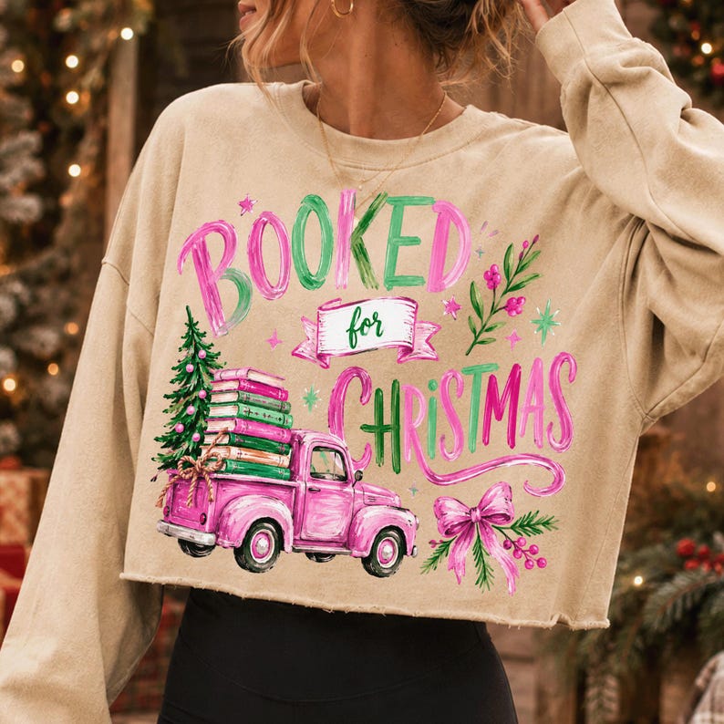 May include: A beige long-sleeve cropped sweatshirt featuring a graphic print with the text "BOOKED for CHRISTMAS". The design includes a pink vintage truck carrying a stack of books and a Christmas tree, accented by a pink bow and greenery.